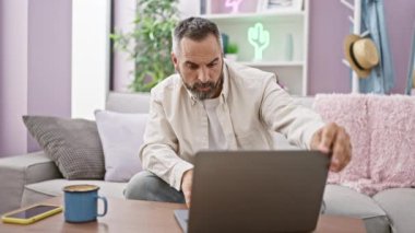 Sakallı ve gri saçlı olgun bir adam ev içinde dizüstü bilgisayarla kanepe ve yastıkla çalışıyor..