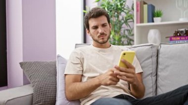 Düşünceli, genç, İspanyol bir adam modern bir oturma odasında oturuyor. Düşünceli bir ifadeyle akıllı bir telefon kullanıyor.