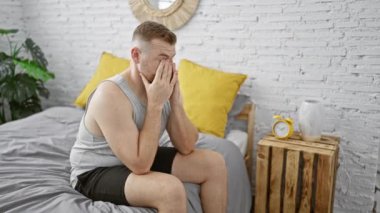 Gündelik kıyafetlerle sakallı bir adam, kapalı bir yatakta oturuyor, sıkıntısını ya da yorgunluğunu ifade ediyor, modern bir yatak odası dekoruyla çevrili..