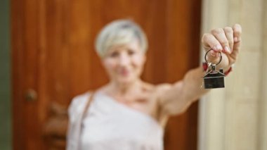 Gri saçlı gülümseyen olgun bir kadın ev anahtarlarını ahşap bir kapının önünde tutuyor. Ev sahipliğini ve yeni başlangıçları simgeliyor..