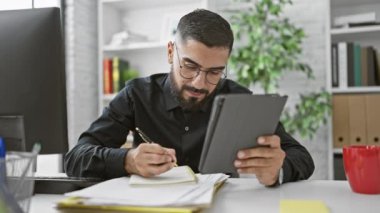 Gözlüklü sakallı bir adam tablet üzerinde çalışıyor ve ofis ortamında notlar alıyor..