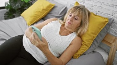 Güzel, orta yaşlı, üzgün bir sarışın kadın, yatak odasında rahat bir şekilde uzanıyor, akıllı telefon kullanımına dalmış. Kapalı bir ortam, rahat bir sabah hissi ve ev içi arka plan..