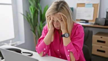 Stresli sarışın kadın modern ofiste bitki ve bilgisayarla yüze dokunuyor.