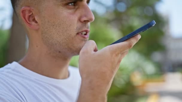 若いヒスパニックの男性は 背景にぼやけた緑の自然を持つ晴れた公園でスマートフォンを使用しています — ストック動画