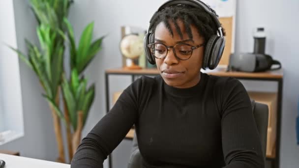 一位快乐的非洲裔美国女人 戴着眼镜和耳机 在现代化的办公环境中工作 — 图库视频影像