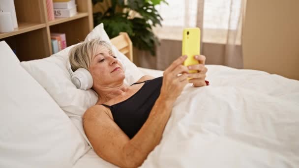 Reife Frau Liegt Mit Kopfhörern Bett Und Benutzt Ein Smartphone — Stockvideo