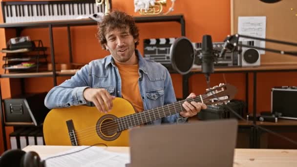 ミュージックスタジオでギターを弾くハンサムなヒスパニック男 マイクと録音機器を備えたラップトップを見る — ストック動画