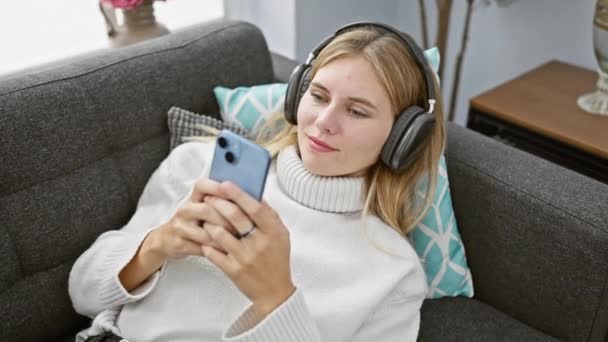 一位金发碧眼的女人带着耳机 在沙发上享受着智能手机的乐趣 在舒适的客厅里传递着闲暇 — 图库视频影像