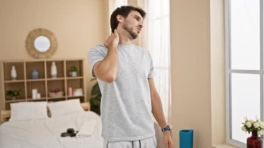 Genç bir adam basit bir dekorla aydınlık bir yatak odasında boynuna masaj yapıyor..