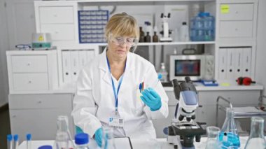 Ciddi, sarışın, orta yaşlı bir bilim adamı, notlar alıyor ve çığır açan tıbbi araştırmalara dalmış, hareketli bir laboratuarda test tüpü tutuyor.