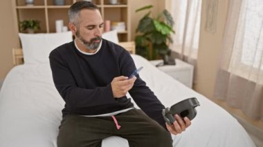 Gri saçlı, akıllı telefon kullanan ve yatak odasında kulaklık tutan olgun sakallı bir adam.