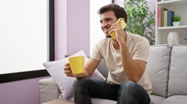 Gülümseyen adam sarı bardağı sıcacık oturma odasında tutarken telefon ediyor..