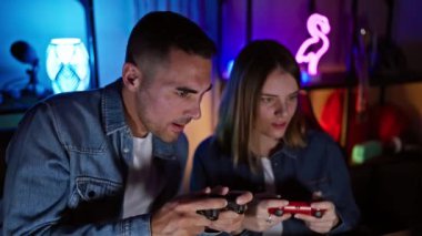 Bir adam ve bir kadın, geceleri neon ışıklı bir oyun odasında yoğun bir şekilde video oyunu oynuyorlar..