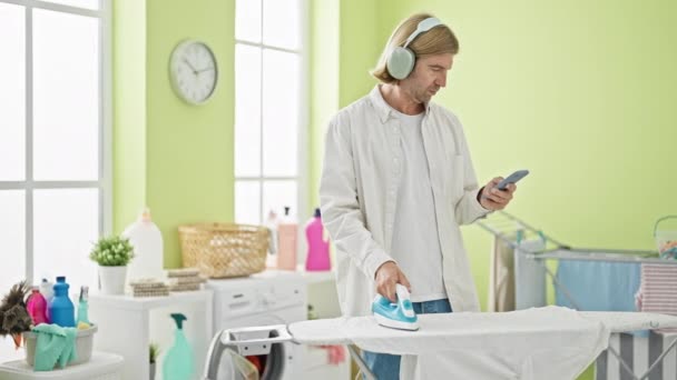 Man Multitasks Ironing Clothes Using Smartphone Headphones Bright Laundry Room — Stock Video