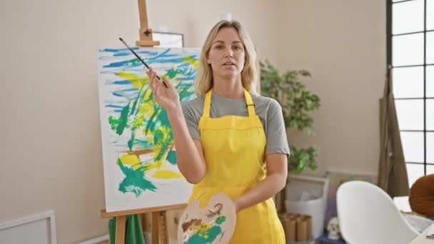 Kontemplacyjna Blondynka Żółtym Fartuchu Stoi Przed Kolorowym Obrazem Jasnym Studio — Wideo stockowe