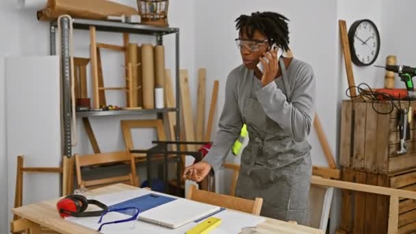 カーペントリーワークショップのアフリカ系アメリカ人女性 電話で 安全メガネと木工設備 — ストック動画