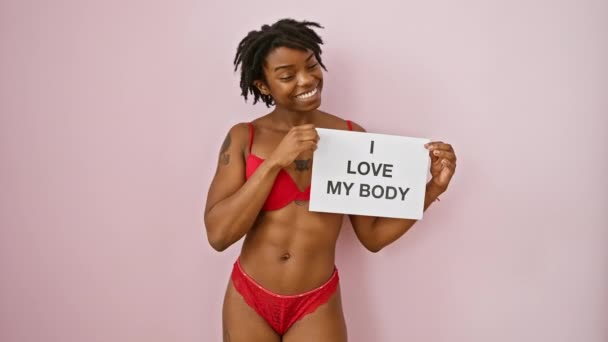 一位穿着红色内衣裤的自信的非洲裔美国女人拿着一个标牌 上面有粉色背景下的身体正面信息 — 图库视频影像