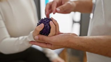 Bir kadın, klinikte erkek bir terapistin yardımıyla dikenli bir topla el terapisi görüyor..