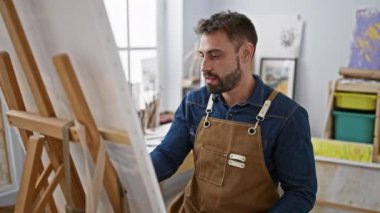 Rahatlamış genç İspanyol bir adam, yakışıklı sakallı bir ressam portresi, resim stüdyosunda fırçalarla resim yapmaya odaklanmış, tuval başyapıtını mükemmelleştiriyor.