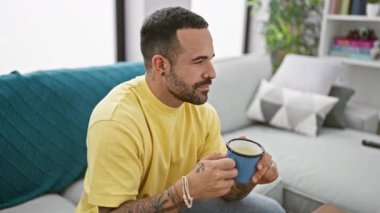 Sarı tişörtlü, dövmeli bir İspanyol evde gevşiyor, modern oturma odasında mavi bir kupa tutuyor..