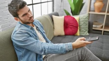 Genç bir İspanyol adam, arka planda modern dekoru olan bir uzaktan kumandayla oturma odasında rahatlıyor..