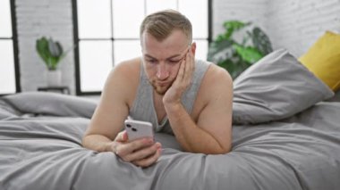 Sakallı dalgın bir genç adam modern bir yatak odası ortamında akıllı telefonunu kullanarak yatakta yatıyor.
