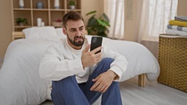 Stresli genç İspanyol bir adam yatak odasında tek başına oturuyor, endişeyle telefonuna mesaj atıyor, sabah baş ağrısı ve endişe ile boğuşuyor.