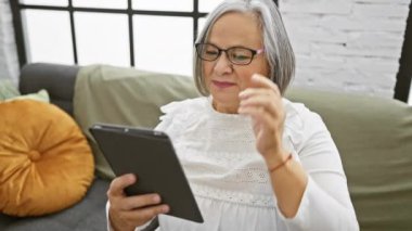 Üst düzey bir kadın güzel ışıklandırılmış bir oturma odasındaki koltukta tablet kullanıyor. Teknoloji, eğlence ve modern yaşam tarzını somutlaştırıyor..