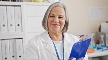 Beyaz laboratuvar önlüklü kendinden emin yaşlı kadın elinde bir dosya tutuyor.