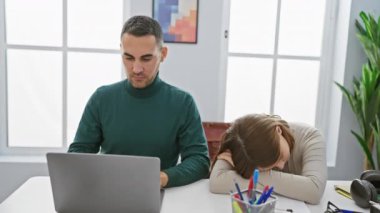Stresli hisseden bir kadın ve modern bir ofiste dizüstü bilgisayarı ve arka planda bitkileri olan bir teselli adamı..