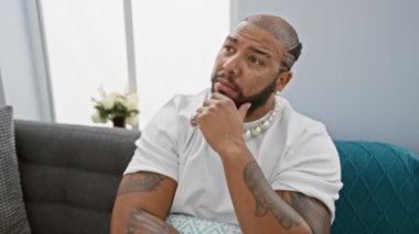 Düşünceli Afrikalı Amerikalı adam evinde oturuyor, eli çenesinde, ciddi bir yüz karışıklığı ile dolu, meydan okuyucu bir fikir üzerinde kafa yoruyor, kanepeye dalmış..