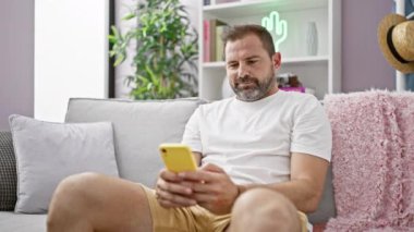 Orta yaşlı, gri saçlı bir adam evdeki bir kanepede akıllı telefon kullanırken gülümsüyor ve sıradan bir yaşam tarzını temsil ediyor..