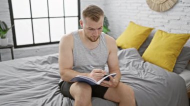 Günlük giysiler içinde sakallı bir adam yatak odasında yastıklarla otururken bir deftere düşünceli bir şekilde yazar..