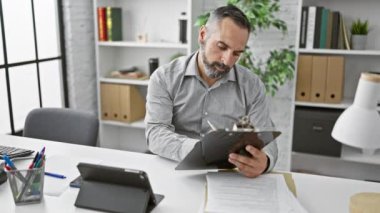 Gri sakallı, olgun bir adam modern ofis ortamında çalışıyor. Pano ve akıllı telefon kullanıyor..