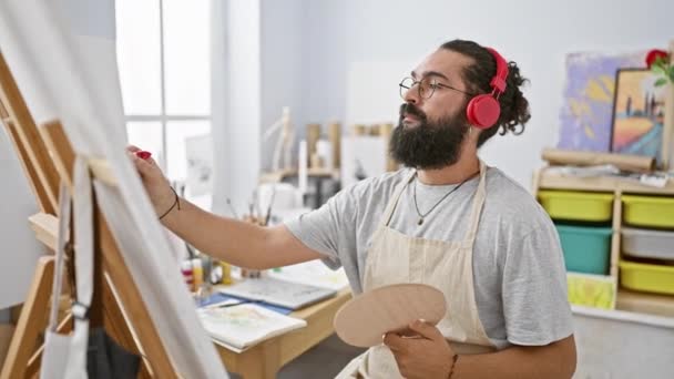 一个留着胡子的男人在明亮的艺术工作室里画画时 一边用红色耳机听音乐 — 图库视频影像