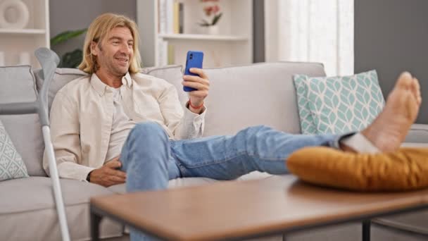 ブロンド男はスマートフォンとのソファーでリラックスし 近代的な居心地の良いリビングルーム — ストック動画