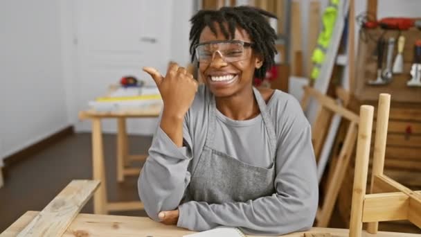 兴高采烈的年轻黑人妇女 留着可怕的头发 戴着安全眼镜 做木工 把大拇指指向旁边 带着张开嘴自信的车间肖像笑着 — 图库视频影像