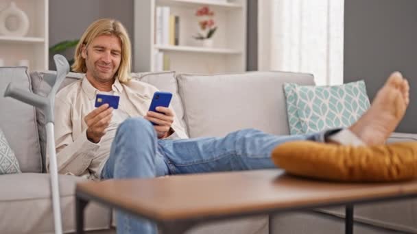 一个悠闲的男人坐在家里的沙发上 看着信用卡和智能手机 旁边夹着拐杖 暗示着一种复苏的情景 — 图库视频影像