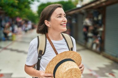 Orta yaşlı kadın turist, elinde şapkayla gülümsüyor.