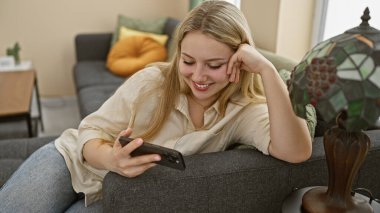 Genç bir kadın evinde bir kanepede dinleniyor, akıllı telefonu tutarken gülümsüyor..