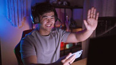 Akıllı telefon kullanan ve neon ışıklı oyun odasında el sallayan, kulaklıklı, gülümseyen Asyalı bir adam.