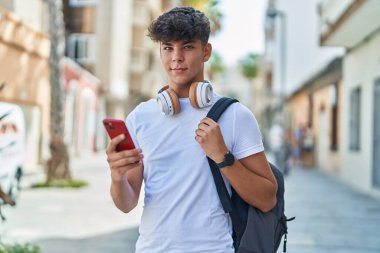 Latin kökenli genç bir öğrenci sokakta akıllı telefon kullanarak gülümsüyor.