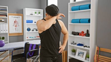 Genç Asyalı bir adam, teçhizatla dolu modern bir rehabilitasyon kliniğinin terapi odasında esniyor..