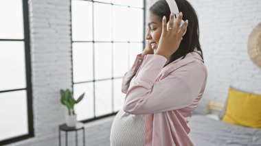 Genç bir kadın için neşe dolu bir sabah, yatakta dinlenen, ezgilere dalmış, göbeğini sallayan ve ışıl ışıl gülümseyen hamile bir kadın için.