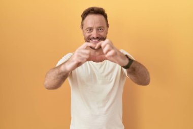 Sarı arka planda dikilen sakallı, aşık bir orta yaşlı adam elleriyle kalp sembolü yapıyor. romantik kavram. 