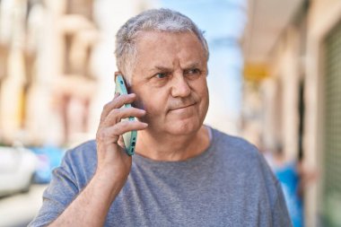 Orta yaşlı, kır saçlı, akıllı telefondan konuşan, sokakta rahat bir yüz ifadesi olan bir adam.