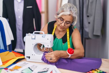 Orta yaşlı, gri saçlı kadın terzi dükkanında dikiş makinesi kullanıyor.
