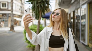 Sarışın kadın güneşli bir caddede selfie çekiyor.