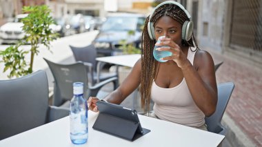 Afro-Amerikalı kadın dokunmatik ped ve kulaklık kullanıyor. Kahve dükkanının terasında bir bardak su içiyor.
