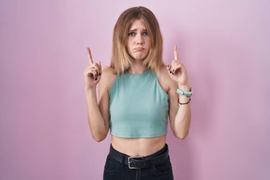 Blonde caucasian woman standing over pink background pointing up looking sad and upset, indicating direction with fingers, unhappy and depressed. 